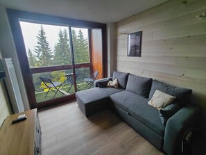 A modern living room with a grey sectional sofa, a small table, and a balcony with a view of trees. - Le Corbier: a new, cozy apartment right in the heart of the resort! (Villarembert)