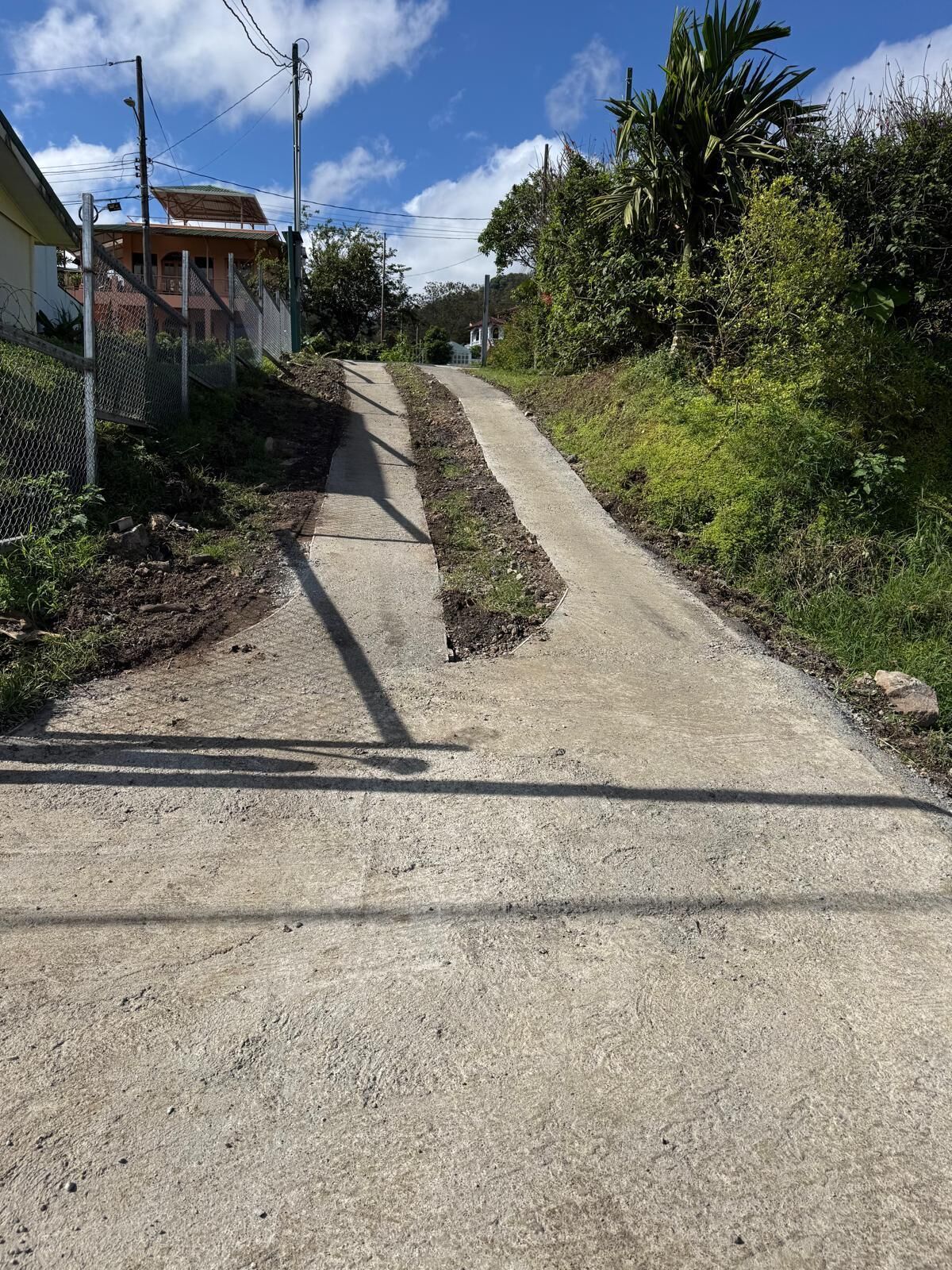 A narrow, unpaved road with a concrete path on one side, surrounded by vegetation and a residential building.