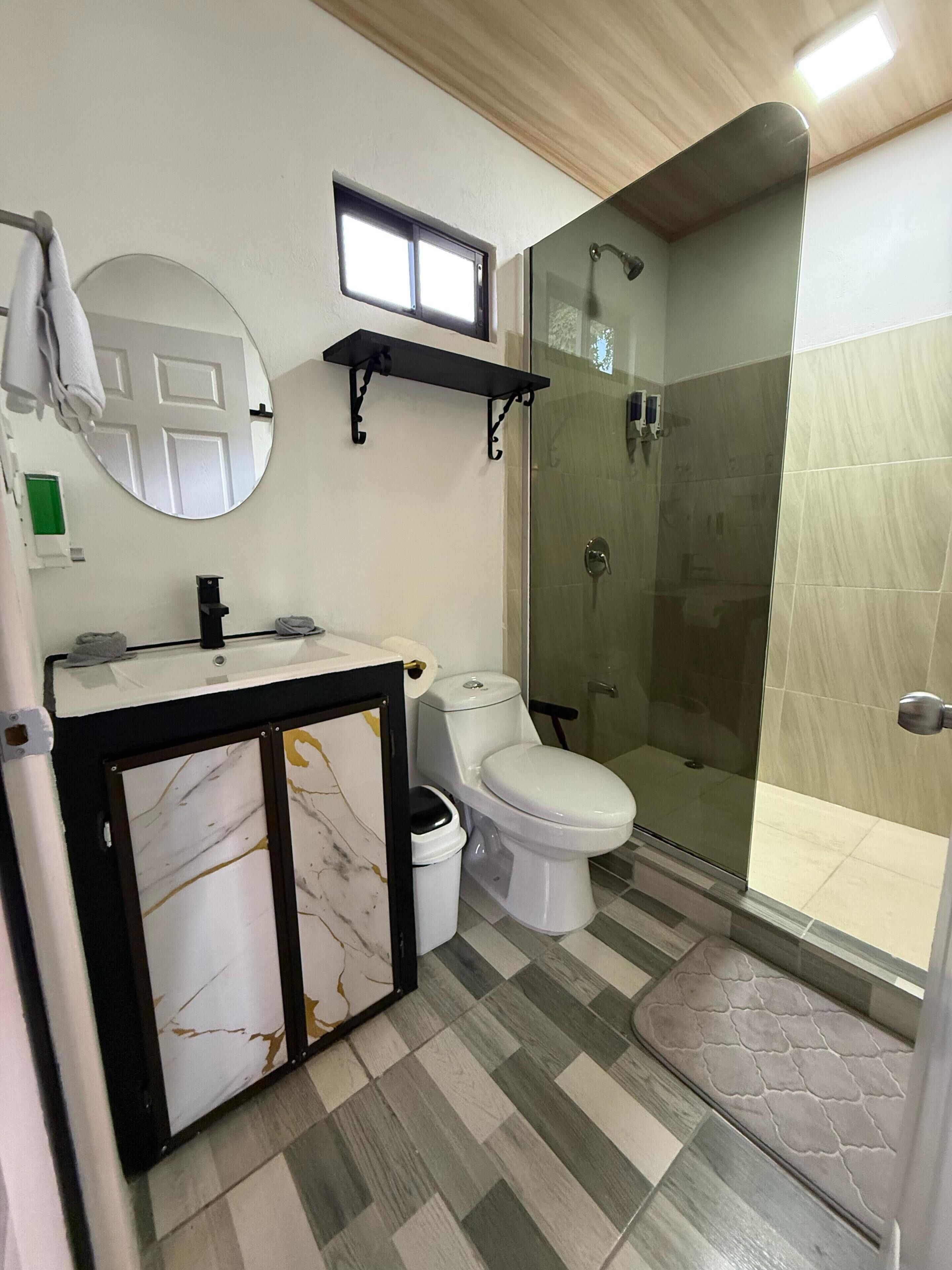 Ein Badezimmer mit Toilette, Waschbecken, Spiegel, Dusche und Fliesenboden.