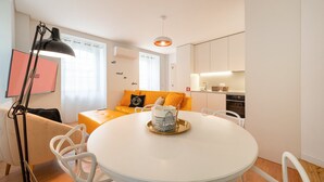Una sala de estar moderna con una mesa redonda blanca, un sofá con cojines anaranjados, una lámpara de pie negra y una cocina con armarios blancos.