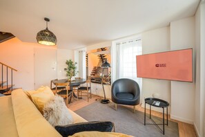 Ein modernes Wohnzimmer mit einer Couch, einem Esstisch, einem Sessel, einer Lampe und einem Fernseher, der 'GUEST READY' anzeigt.