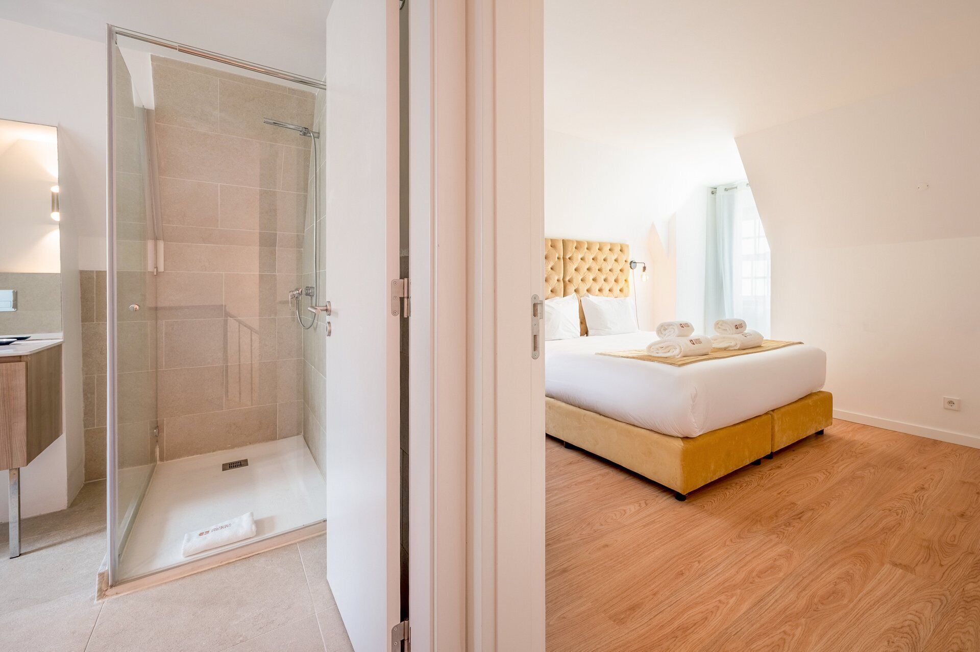 Quarto de hotel com chuveiro em cabine de vidro, cama com cabeceira de madeira e piso de madeira.