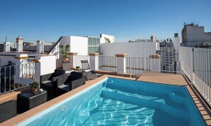 Una piscina en la azotea con un cielo azul despejado, paredes blancas y algunas plantas en macetas.
