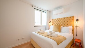 A hotel room with a bed, a bedside table, a nightstand, a window, and an air conditioning unit. - Guestready - The Corner 2D (Lisbon)
