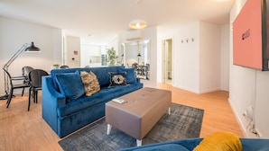 Ein modernes Wohnzimmer mit einem blauen Sofa, einem grauen Hocker und einem Essbereich mit einem schwarzen Tisch und Stühlen.