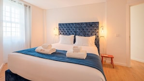 Ein Hotelzimmer mit einem großen Bett, einem blauen Kopfteil, weißen Kissen und einer blauen Bettdecke. Zwei weiße Handtücher mit einem Logo sowie ein kleiner Holzschemel befinden sich neben dem Bett.
