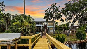 A wooden deck leading to a house with a metal roof, surrounded by tropical vegetation and a body of water. - Fisherman's Cove -Waterfront home with private dock on the River sleeps 10 (Steinhatchee)