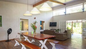Amplia sala de estar con una mesa de comedor de madera, bancos y un sofá. Hay lámparas colgantes, un televisor y vistas al exterior.