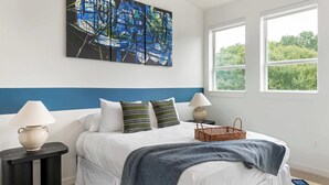 Una camera da letto con un letto, due comodini con lampade, un quadro astratto di grandi dimensioni, una finestra vista alberi e un tappeto decorato.
