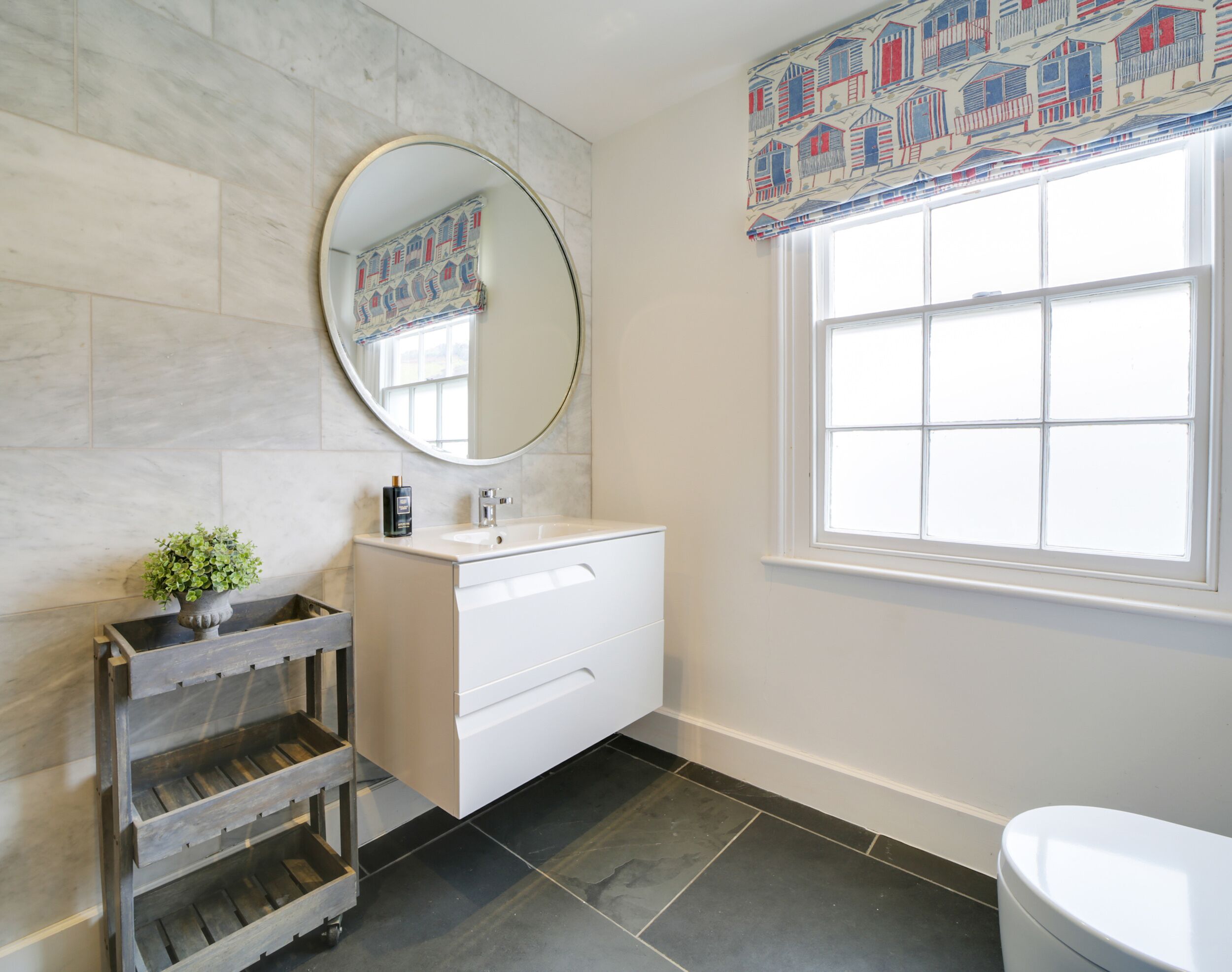 A bathroom with a white vanity, a round mirror, a potted plant, and a window with a patterned valance.