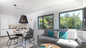 Un soggiorno moderno con zona pranzo, un divano grigio con cuscini color petrolio e un'ampia finestra che si affaccia su alberi e una scala.