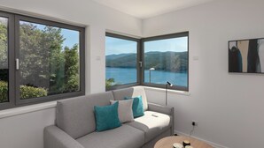 Un soggiorno moderno con un divano grigio, cuscini color petrolio e vista su un lago e montagne attraverso ampie finestre.