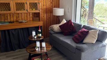 Ein Zimmer mit einer Holzwand, einem grauen Sofa, einem kleinen runden Tisch und einem Fenster mit Blick auf Grünfläche.