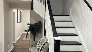 Un escalier avec des rampes noires et des marches blanches mène à l’étage supérieur. Un banc agrémenté d’un coussin à motifs est placé au pied de l’escalier.