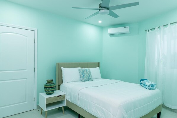 Un dormitorio con cama, mesita de noche, ventilador de techo, equipo de aire acondicionado y una puerta.