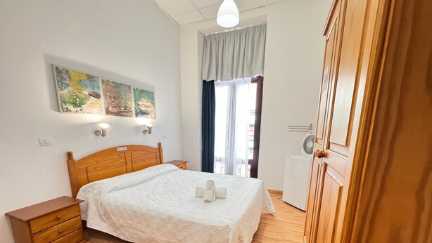 Un dormitorio ordenado con una cama de madera, una mesita de noche, una lámpara de pared y un ventanal con cortinas.