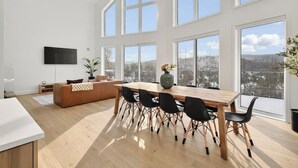 Een ruime woonkamer met een grote houten eettafel, zwarte stoelen, een bank en een aan de muur bevestigde televisie.