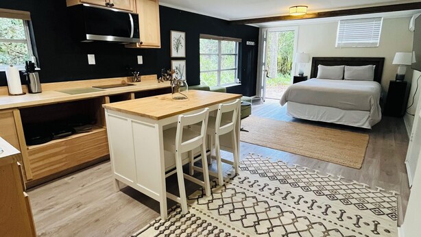 Una cocina compacta y moderna con armarios de madera y una isla central, un dormitorio acogedor con una cama grande y mesitas de noche, y una alfombra estampada en el suelo.