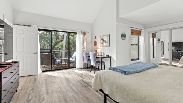 Ein geräumiges Schlafzimmer mit einem großen Bett, einem Schreibtisch und einem Sessel, ein Balkon mit Blick auf Bäume und eine Schiebetür zu einem weiteren Raum.