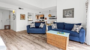 Ein modernes Wohnzimmer mit einem blauen Sofa, einem Korbtisch und einer Küche im Hintergrund.