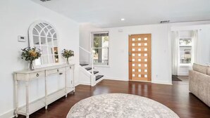 Un salón amplio con suelo de madera, una consola blanca con jarrones, una escalera y una puerta de madera.
