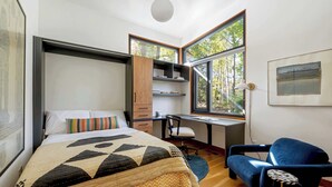 Ein modernes Schlafzimmer mit Bett, Schreibtisch und Stuhl, großen Fenstern und Blick auf Bäume.