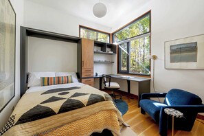 A modern bedroom with a bed, desk, and chair, large windows, and a view of trees. - Glass Grove (Atlanta)