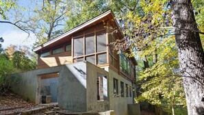 Ein modernes, zweistöckiges Haus mit großen Fenstern und Holzfassade, umgeben von Bäumen und einem Betonfundament.