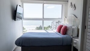 A bedroom with a large window, a bed with blue bedding, a white nightstand, and a TV mounted on the wall. - Ventanas Ocean View -V3-7C- 2 bed, 2 bath (Punta Chame)