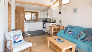 Un salon chaleureux avec un plancher en bois, un canapé bleu agrémenté de coussins décoratifs, une table basse en bois et un fauteuil avec un coussin à motif de phare. On aperçoit, au fond, une cuisine avec des armoires blanches et une fenêtre.