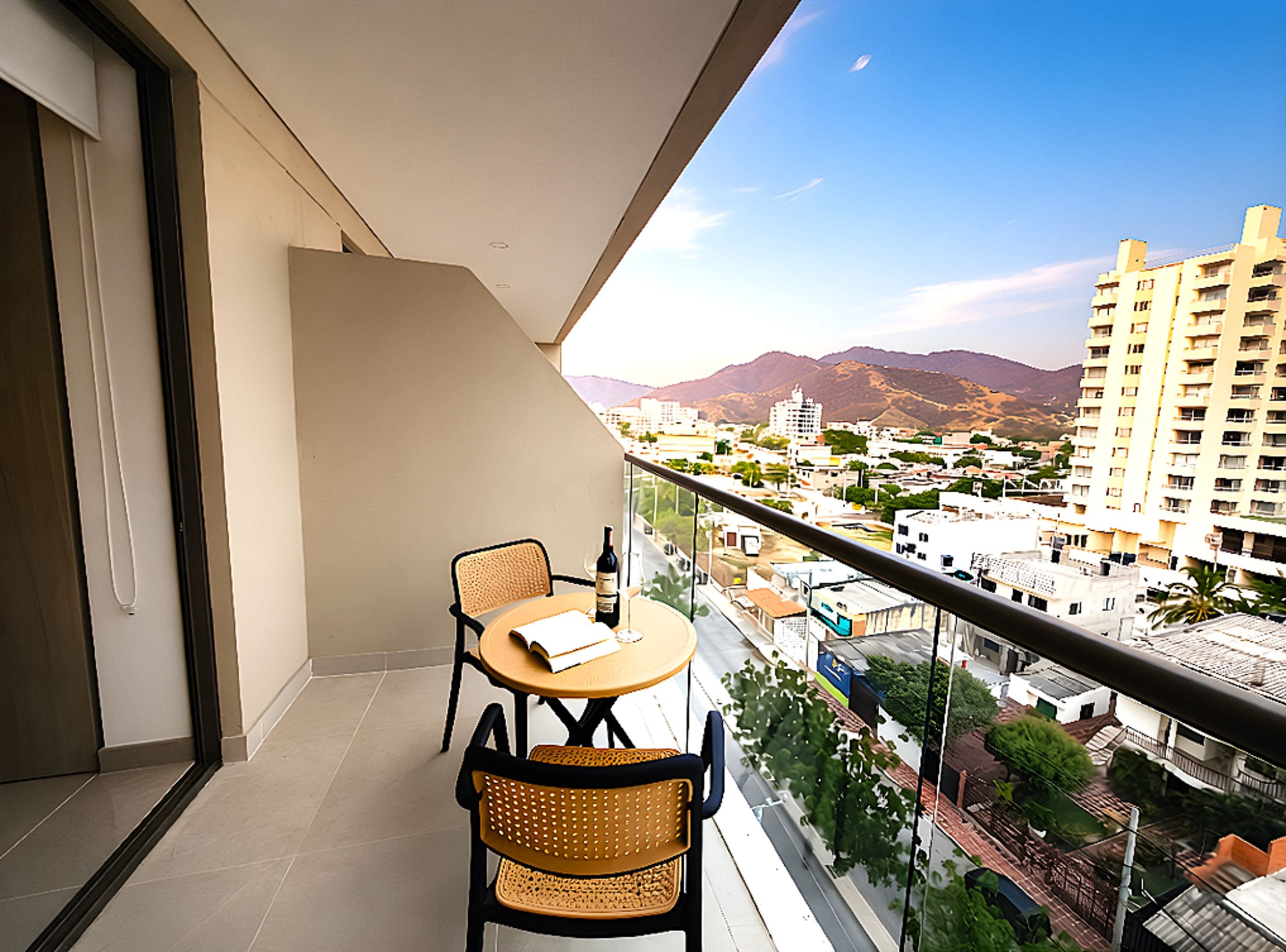 A balcony with a table and chairs, overlooking a cityscape with buildings and mountains.