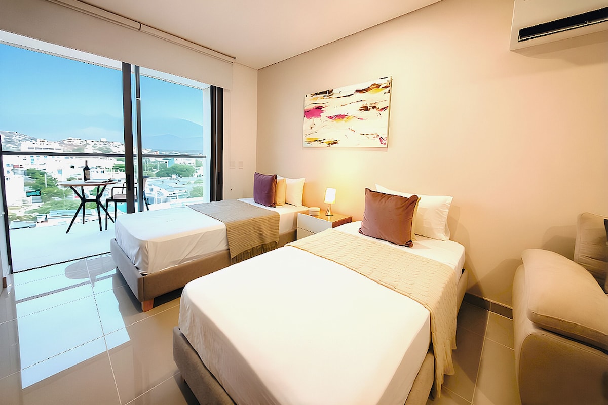 A hotel room with two beds, a balcony, a table with a bottle, and a city view.
