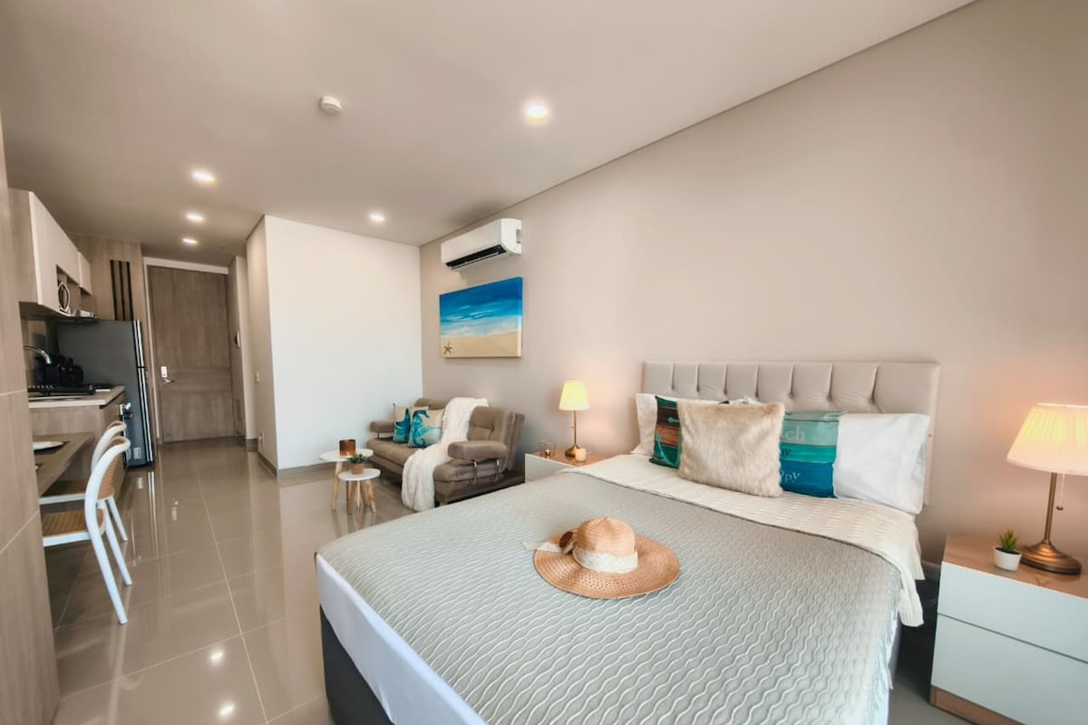 A modern hotel room with a large bed, a small seating area, a kitchenette, and a view of the sea.