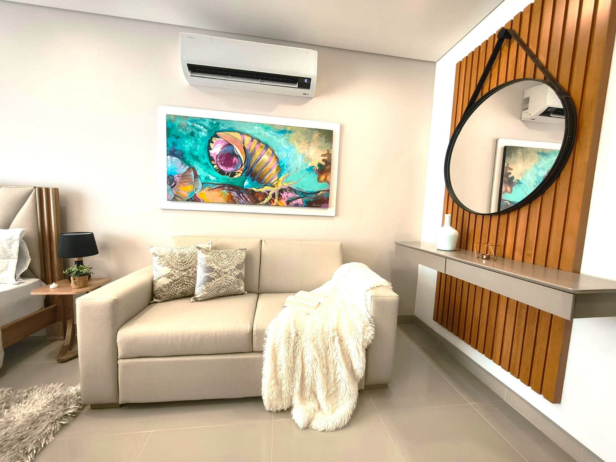 A modern living room with a beige sofa, a fluffy throw blanket, a painting, a round mirror, and a wooden panel wall.