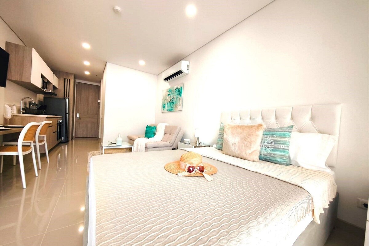 A modern hotel room with a large bed, a dining area with a table and chairs, and a kitchenette.