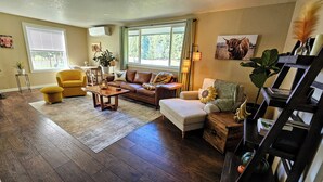 A spacious living room with a brown leather sofa, a yellow armchair, and a wooden coffee table. There is a large window providing a view of the outdoors. - Private Ranch House, Highland Cattle, Horses & Peaceful Views - a soul refresher (Myrtle Creek)