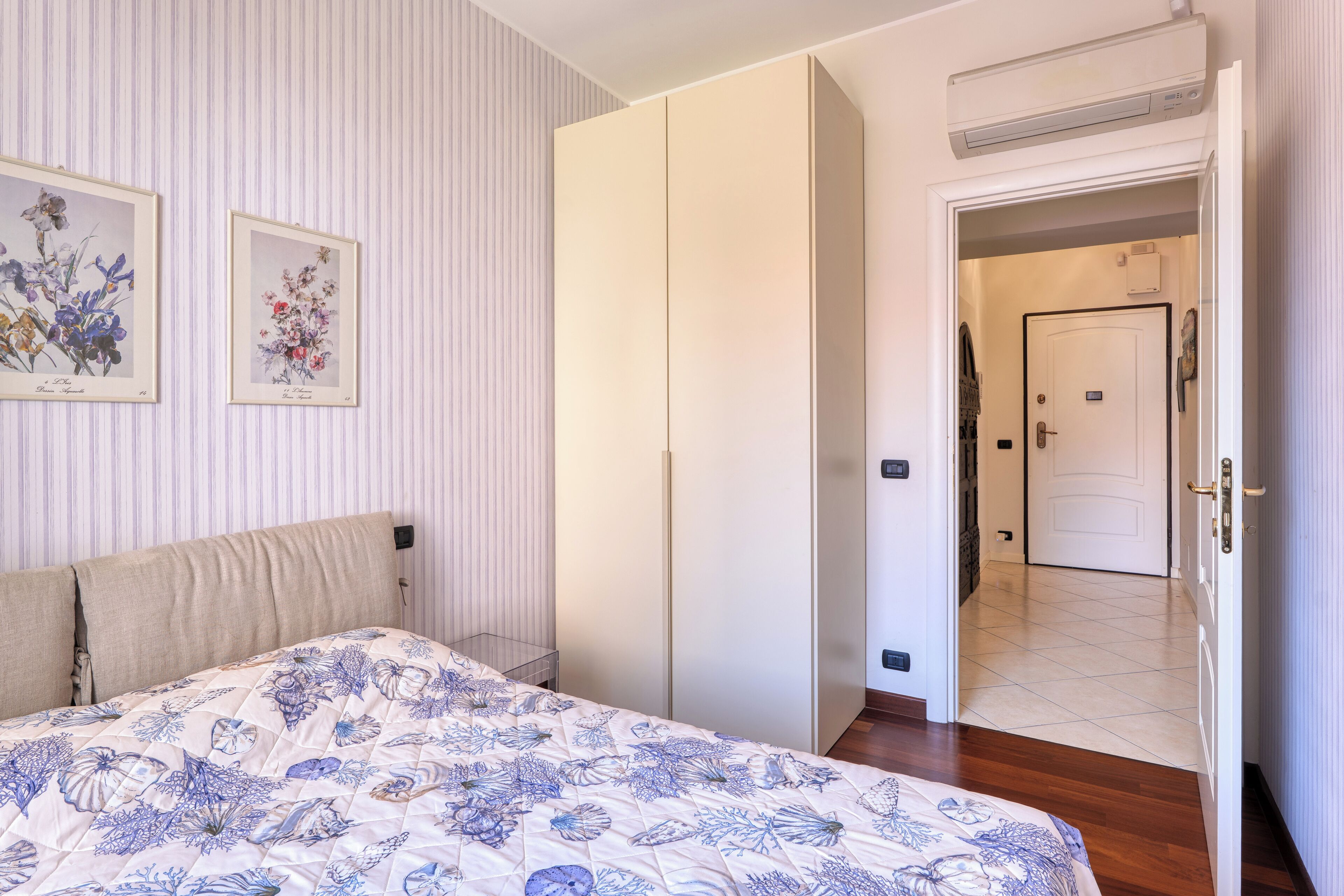 A bedroom with a bed, a wardrobe, a door leading to another room, and a framed floral artwork on the wall.