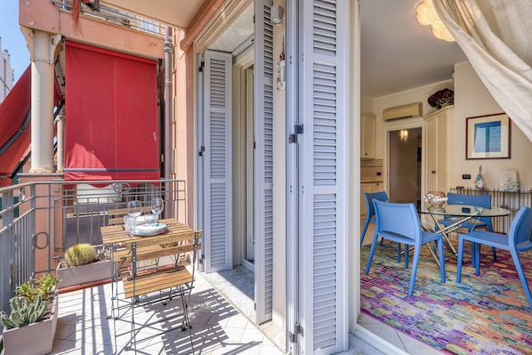 A balcony with a red awning, a table with chairs, and a colorful rug inside a room. - Palm House Apartment in an elegant, central setting, just a few steps from the ocean. (Bordighera)