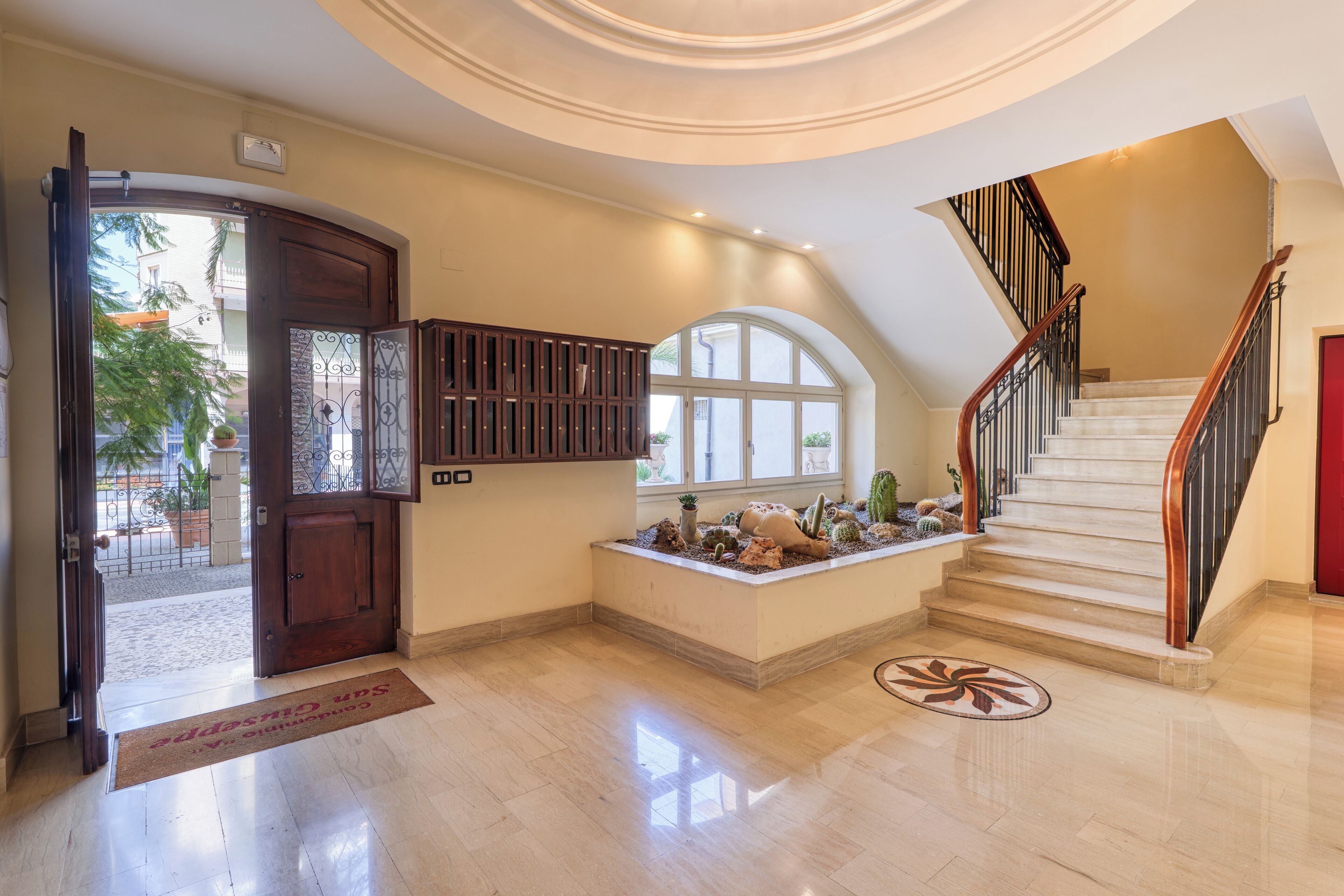 A spacious foyer with a polished marble floor, a wooden door, a built-in cabinet, a small cactus garden, and a staircase with a wooden handrail.