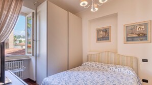 Een slaapkamer met een bed, een raam met gordijnen, een radiator, en twee foto’s in lijsten aan de muur.