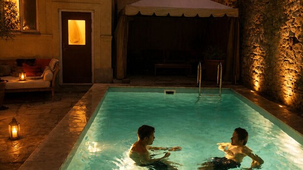 Una piscina rectangular con dos personas nadando, un área de estar techada y una pared de piedra.