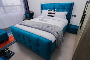 A modern bedroom with a tufted blue headboard, a grey and white bedspread, and a striped pillow. - Padmore Residences Anno (Nairobi)