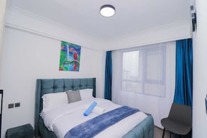 A hotel room with a bed, a chair, a painting on the wall, and a window with curtains. - Padmore Residences Anno (Nairobi)