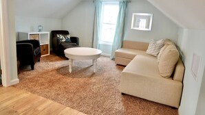 Ein gemütliches Wohnzimmer mit einem beigen Sofa, einem schwarzen Sessel, einem weißen Couchtisch und einer kleinen Kommode.