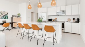 Una cocina moderna con armarios blancos, una isla central con taburetes de bar, iluminación colgante y un refrigerador a la vista.