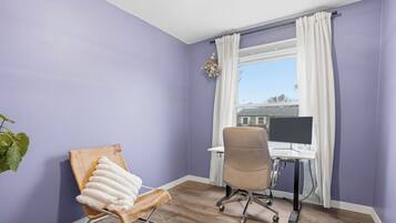Una habitación con pared morada, suelo de madera, un escritorio con computadora, una silla con cojín blanco y una ventana con cortinas.