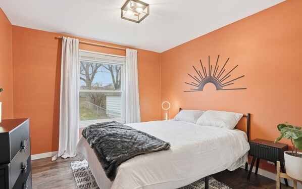 A bedroom with a bed, a window with curtains, a wall decoration, a bedside table, and a potted plant.