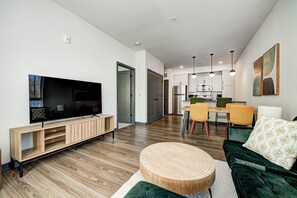A modern living room with a flat-screen TV, wooden entertainment unit, green sofa, round wooden coffee table, and a dining area with yellow chairs and a wooden table.