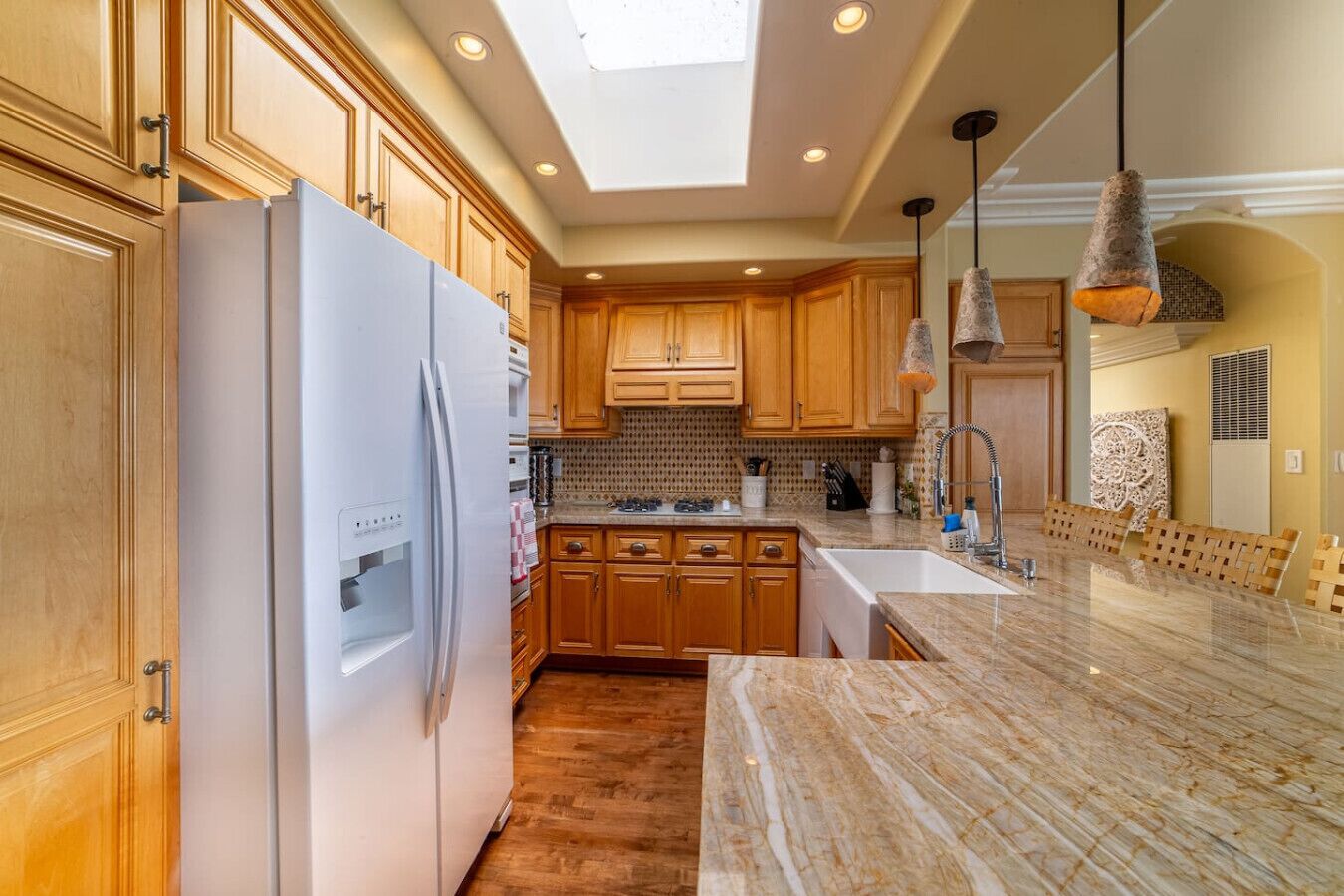 Cucina con mobili in legno, frigorifero bianco, piano di lavoro in marmo e illuminazione a sospensione.