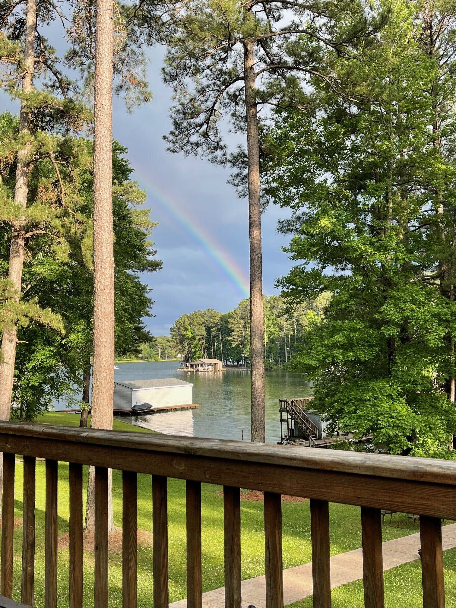 A wooden deck overlooking a lake with a rainbow, surrounded by trees and a dock with a building.